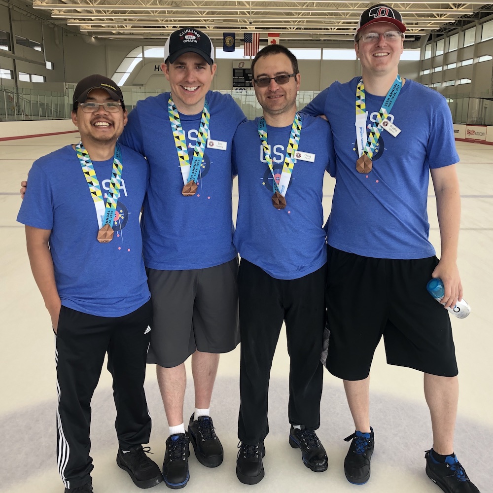 Photo of Adam Leong (left) standing with his "Double for Five" teammates at the Cornhusker State Games. All four curlers are wearing blue t-shirts and bronze medals around their necks.